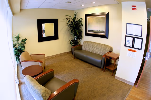 A small waiting area with a patterned carpet, two armchairs, a small round wooden table, a two-seater sofa, a side table, a large potted plant, a framed mirror, and a framed painting on the walls. There are also some framed certificates and a warning sign on the wall near an open door.