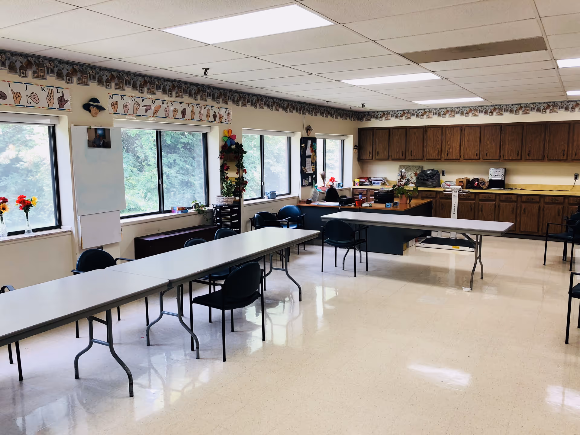 Spacious multipurpose dining/activity room with long folding tables, chairs, windows, and a kitchenette with wood cabinets.
