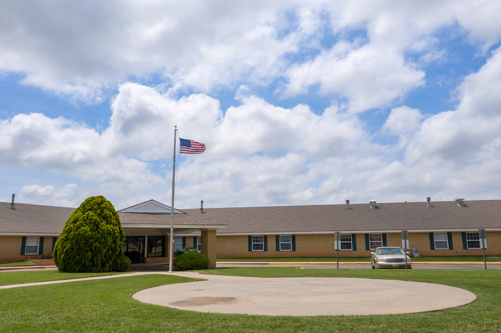 Exterior view of a single-story assisted living facility building with a large green lawn in front, an American flag on a flagpole, a circular concrete area, and a parked car near the entrance under a partly cloudy sky.