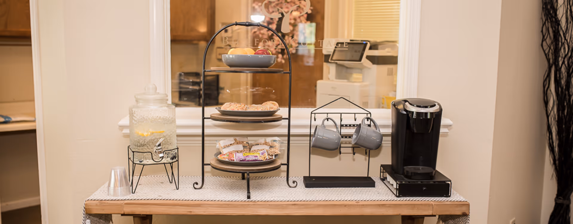 A refreshment station on a wooden table with a glass dispenser filled with water and lemon slices, a three-tiered tray holding fruit, bagels, and packaged snacks, two hanging gray mugs on a metal rack, and a black single-serve coffee maker. The background shows a window into another room with wooden cabinets and a printer.