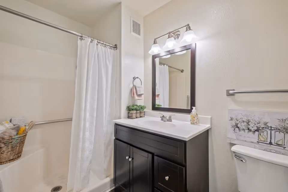A clean bathroom with a shower area featuring a white curtain and a grab bar, a dark wood vanity with a white countertop and sink, a mirror with three light fixtures above it, a small plant decoration, a soap dispenser, a towel ring with folded towels, and a toilet with a floral picture above the tank.