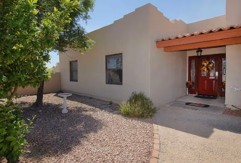Exterior view of a southwestern-style building with beige stucco walls, a red front door decorated with a wreath, a small garden area with gravel, a birdbath, and some greenery including a tree and bushes under a clear blue sky.