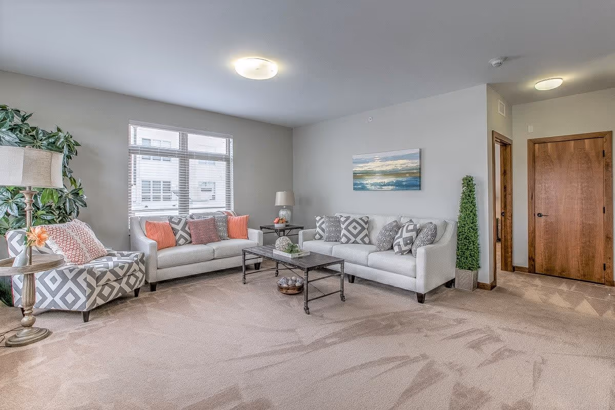 A bright living room with two white sofas adorned with patterned and solid coral and gray pillows, a patterned armchair with a coral pillow, a wooden coffee table with decorative items, a floor lamp, a large window with blinds, a potted plant, a painting on the wall, and a wooden door.