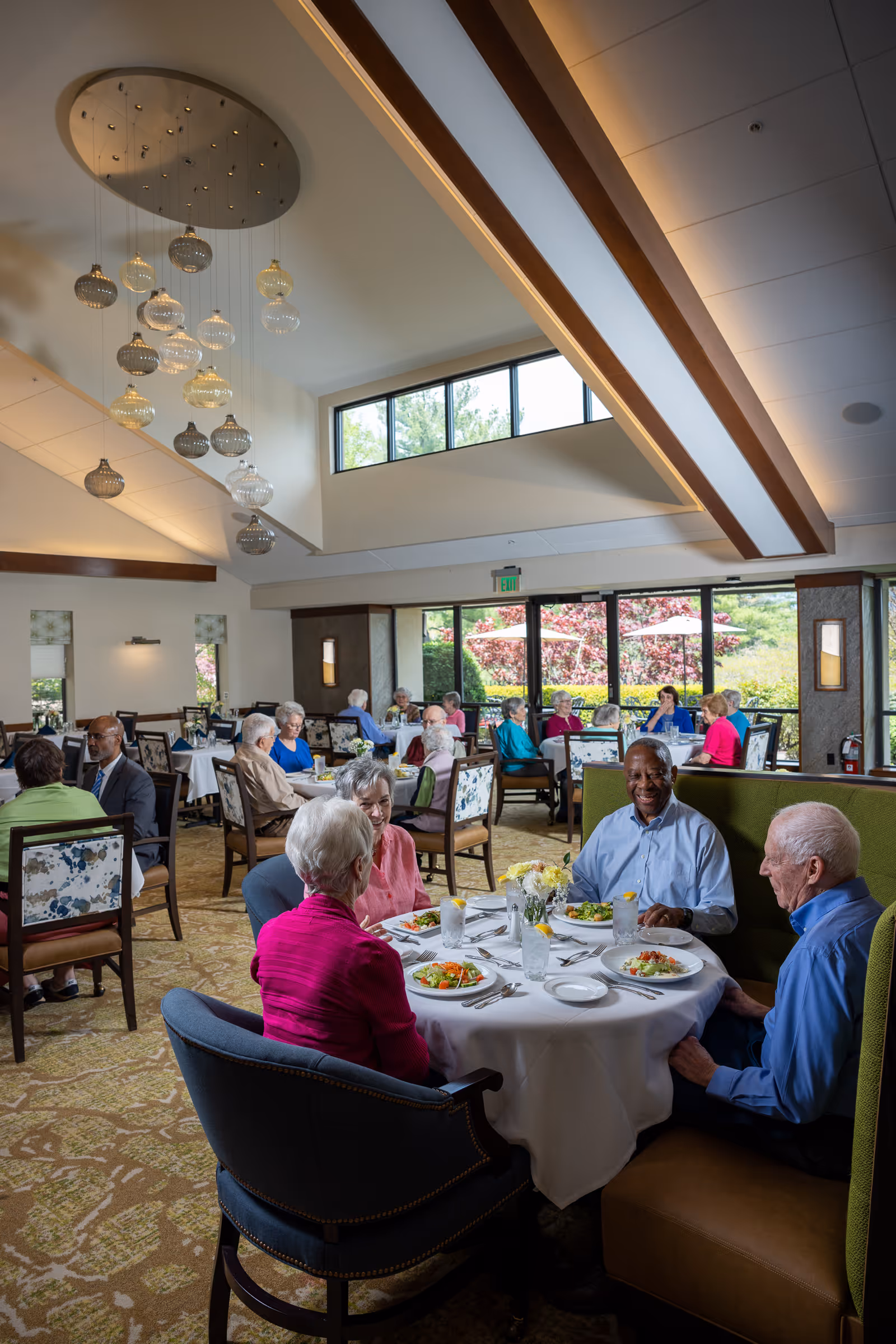 A group of elderly people sitting around a round dining table in a well-lit dining room with large windows showing a garden outside. The table is set with plates of salad, glasses of water, and silverware. Other elderly people are seated at tables in the background, enjoying their meals. The room has a high ceiling with modern hanging light fixtures and a carpeted floor.