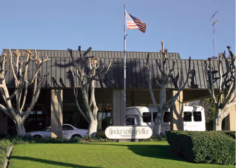 Front entrance of Linda Valley Villa showing the building facade, flagpole, trimmed trees, lawn, and parked vehicles.