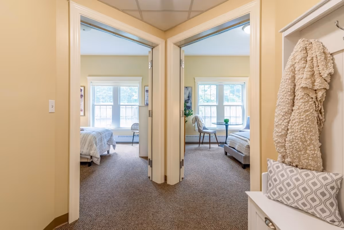 Hallway opening into two sunlit bedrooms with beds and seating by large windows, and a coat and cushion on a bench in the foreground.
