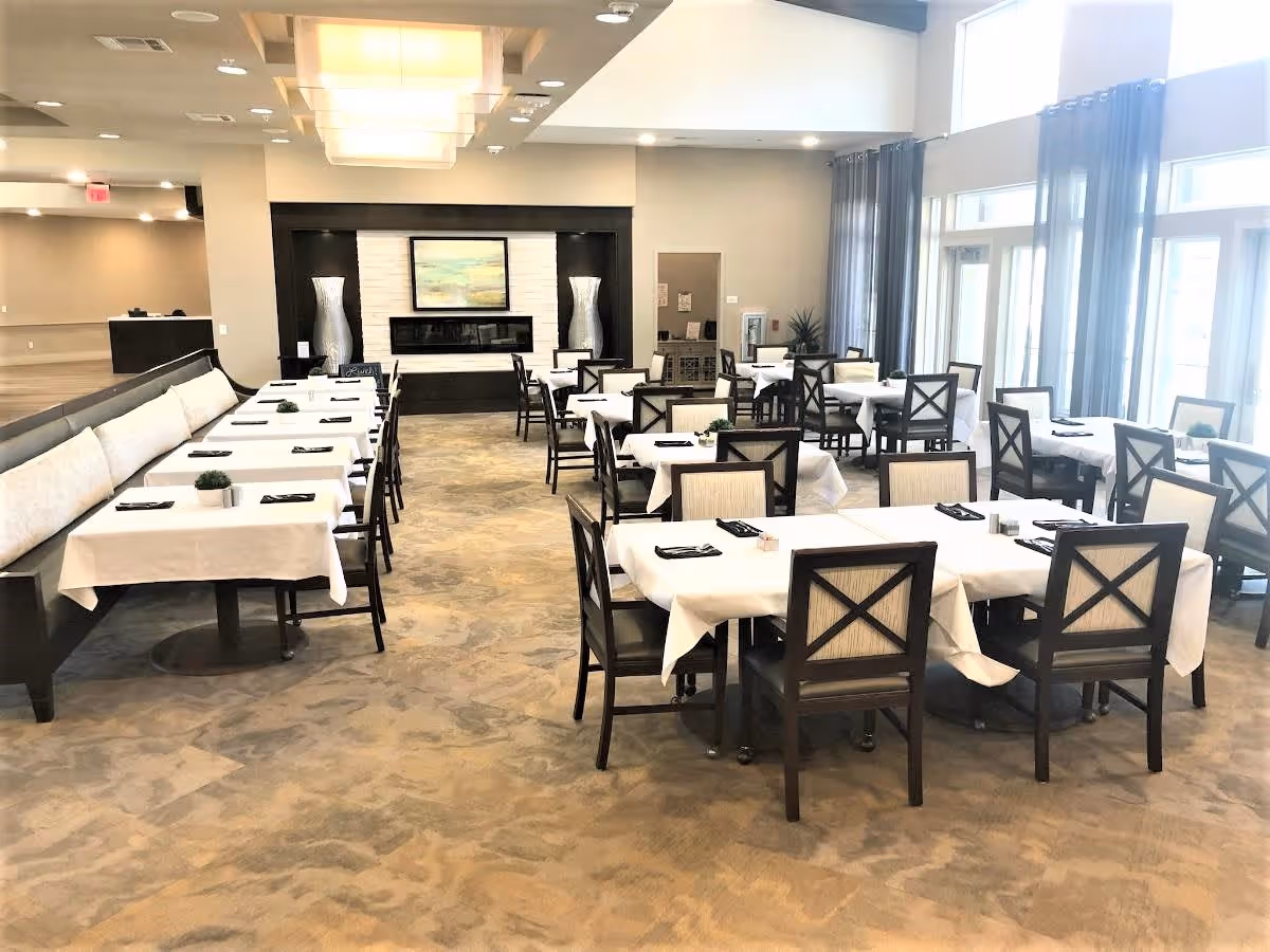 A spacious dining room with multiple tables covered in white tablecloths, each set with black napkins and small decorative plants. The room features large windows with sheer curtains allowing natural light to fill the space. At the far end, there is a modern fireplace flanked by two tall decorative vases and a framed artwork above it.