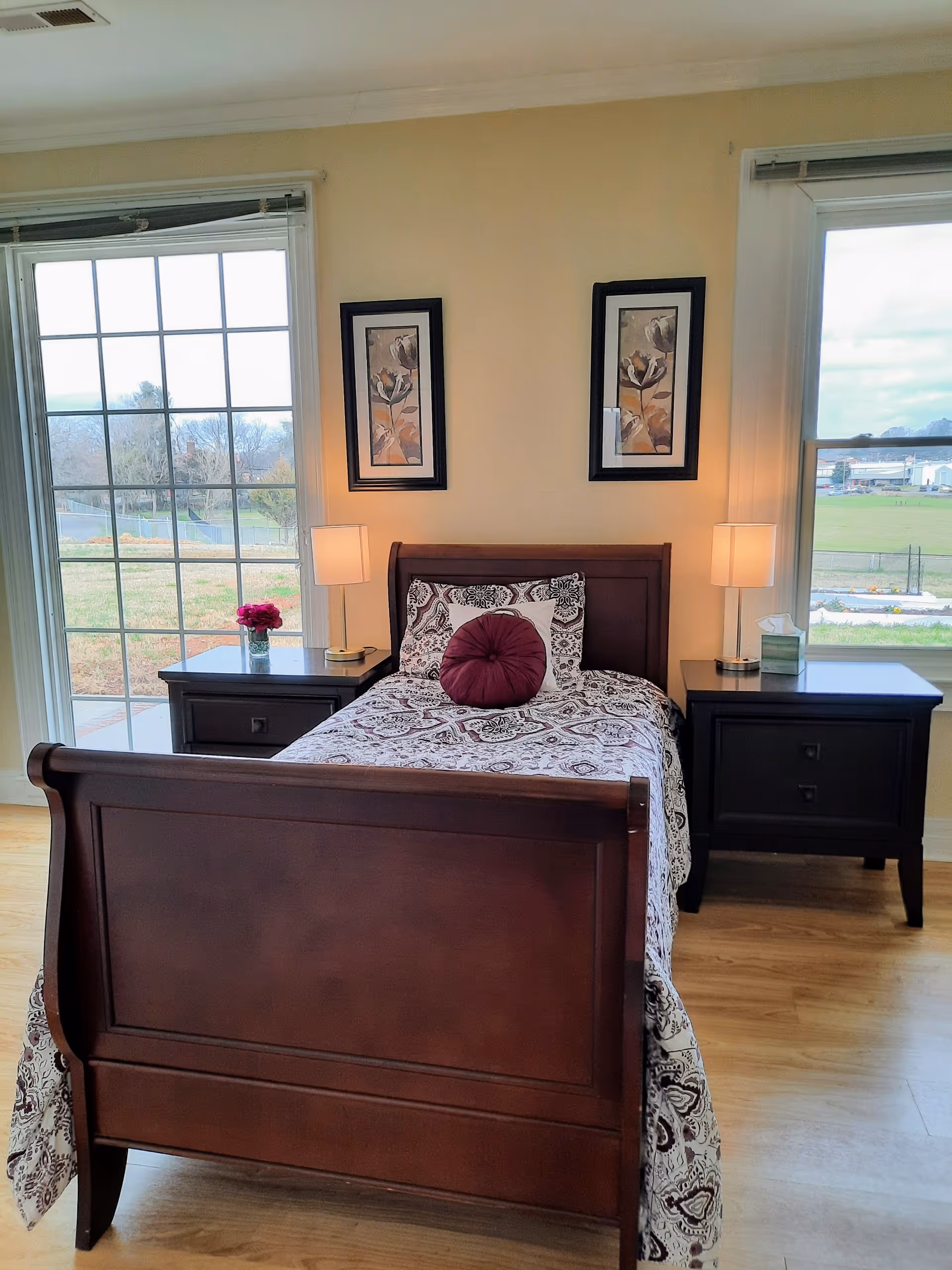 A bedroom with a wooden bed frame and a patterned bedspread, featuring a round purple pillow and a square pillow. There are two matching dark wooden nightstands on either side of the bed, each with a lamp. Two framed floral artworks hang on the beige wall above the bed. Large windows on both sides of the bed provide a view of an outdoor grassy area and buildings in the distance.