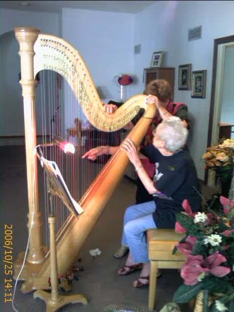 An elderly woman seated on a chair playing a large wooden harp with the assistance of another woman standing behind her in a room decorated with framed pictures and a floral arrangement.
