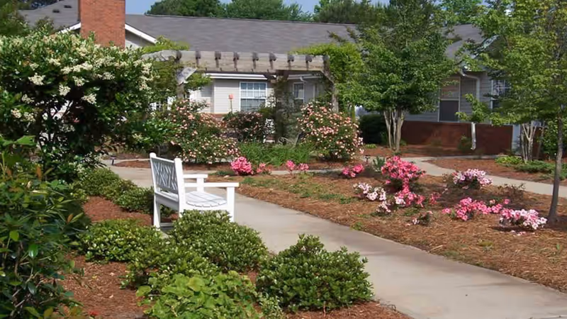 A garden area at Gaines Park Senior Living featuring a concrete pathway, green shrubs, blooming pink flowers, a white wooden bench, and a pergola attached to a building in the background under a clear blue sky.