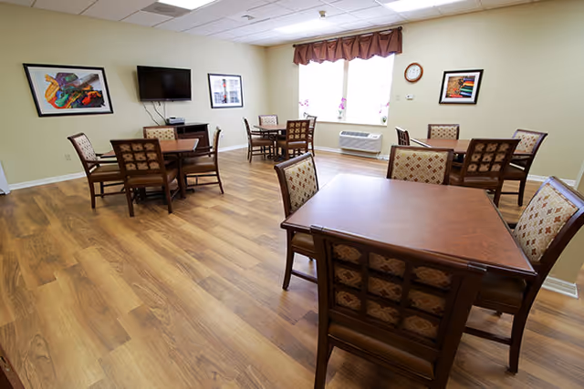 A well-lit common room with wooden flooring featuring several square wooden tables surrounded by cushioned chairs with patterned upholstery. The walls are light-colored and decorated with framed artwork. A flat-screen TV is mounted on one wall above a small cabinet. A window with a brown valance allows natural light into the room.