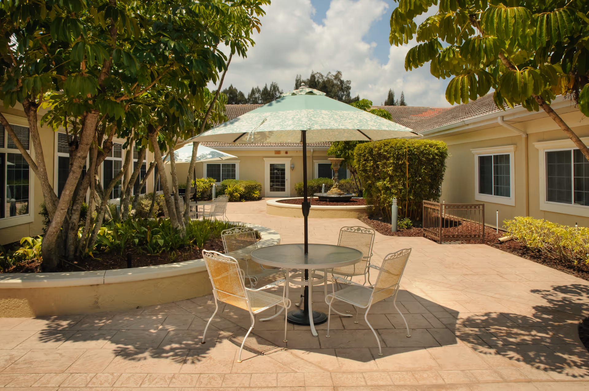 Outdoor courtyard area at Life Care Center of Port St. Lucie featuring a round table with four metal chairs and a large umbrella. The courtyard is surrounded by a single-story building with windows and landscaped with trees, bushes, and a central fountain. The sky is partly cloudy.