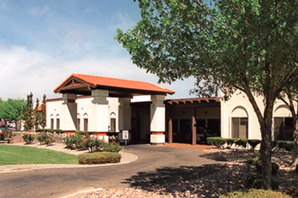 Exterior view of a single-story building with white walls and a red-tiled roof, surrounded by trees, bushes, and a paved driveway under a clear blue sky.