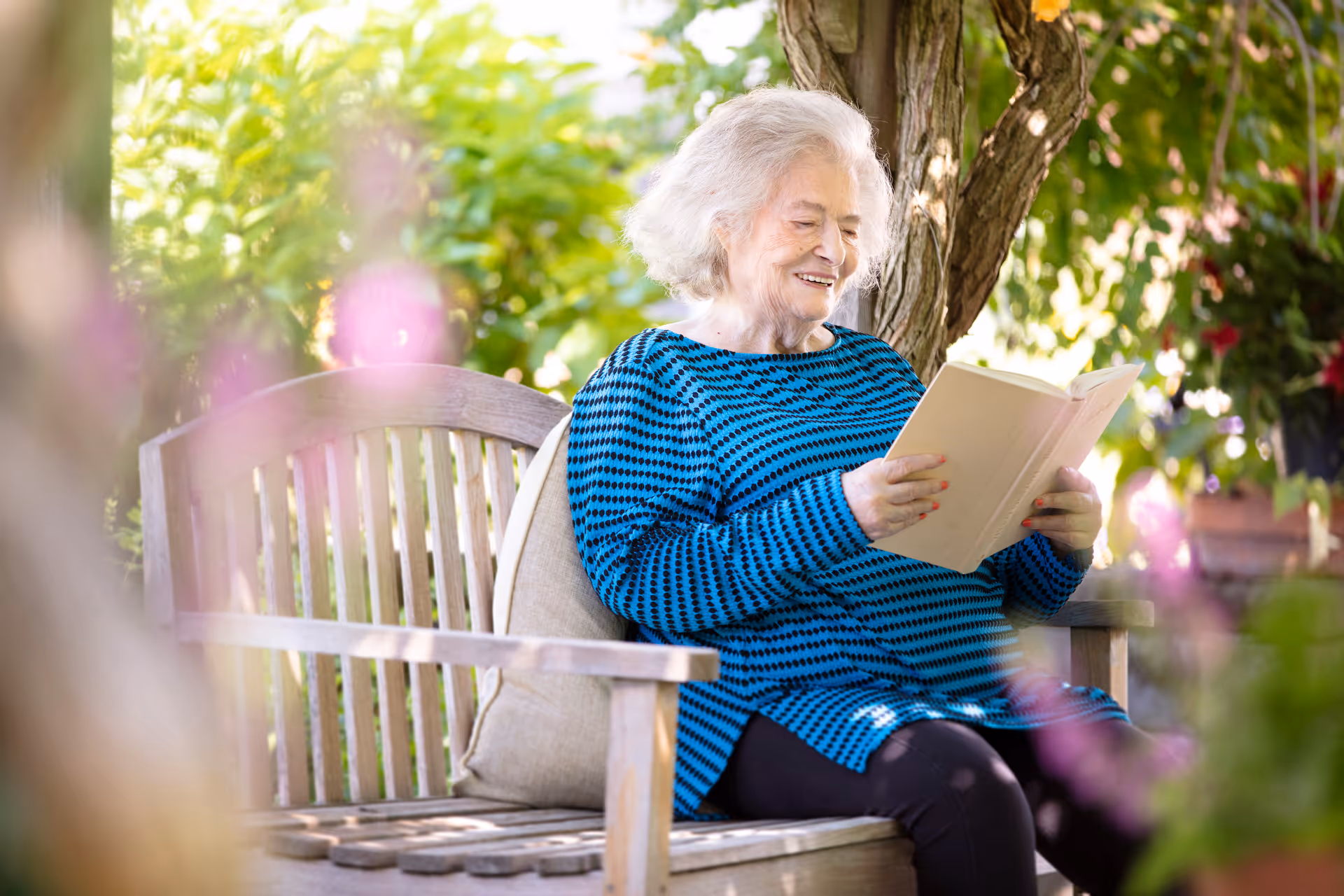 An elderly woman with white hair wearing a blue and black striped sweater is sitting on a wooden bench outdoors, reading a book and smiling. The background is filled with green foliage and flowers, creating a peaceful garden setting.