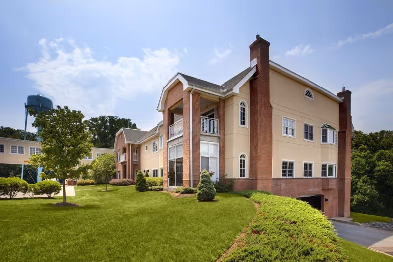 Exterior view of a multi-story senior living building with brick and beige siding, a manicured lawn, and a water tower in the background.