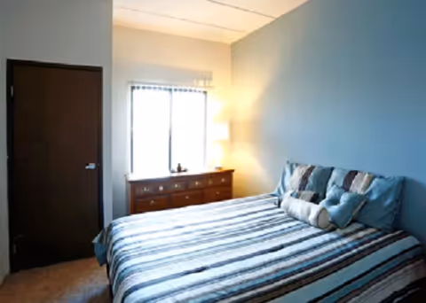 A bedroom with a bed covered in a striped blue, white, and gray comforter. There are several pillows on the bed. A wooden dresser with a lamp and decorative items is positioned against the wall under a window with blinds. A closed dark wooden door is visible on the left side of the room.