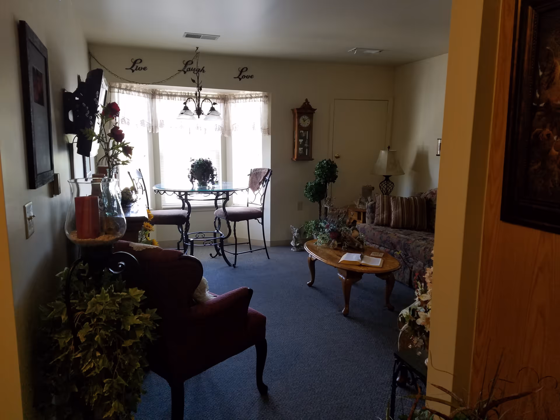 A cozy living room area with a floral patterned sofa, a wooden coffee table with an open book, a standing lamp, and decorative plants. There is a small round glass table with two chairs near a window with sheer curtains. The wall above the window has the words 'Live Laugh Love' displayed. A wooden wall clock is mounted on the wall.