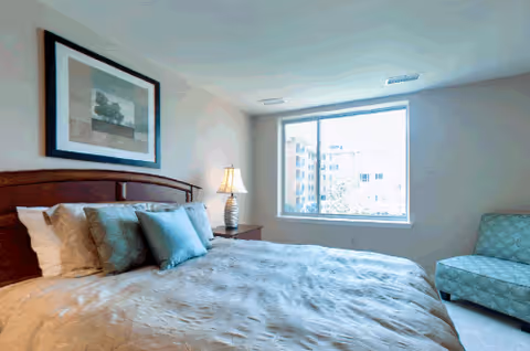 A bright bedroom with a large bed featuring multiple pillows, a wooden headboard, a bedside table with a lamp, a framed picture on the wall, a large window letting in natural light, and a patterned armchair in the corner.