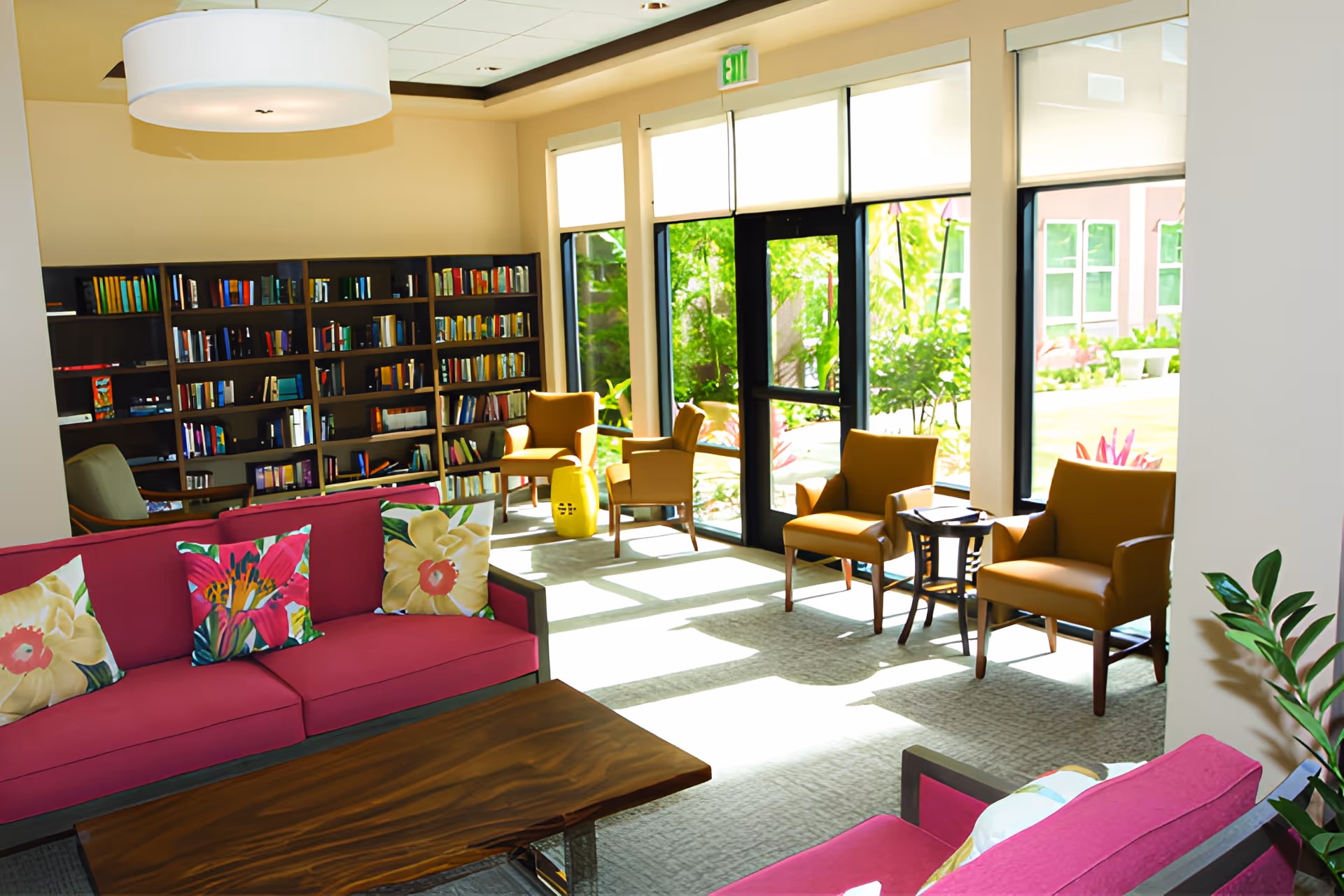 Sunlit community lounge with bookshelves, pink sofas, armchairs, and large windows overlooking a garden.