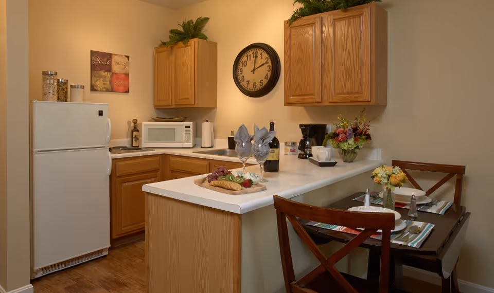 Small kitchen and breakfast bar with oak cabinets, white appliances, a bottle of wine and glasses on the counter, and a two-person dining table set.