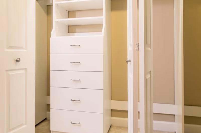 A white built-in closet organizer with four drawers and two open shelves at the top, situated between two open doors in a room with beige walls and tiled floor.