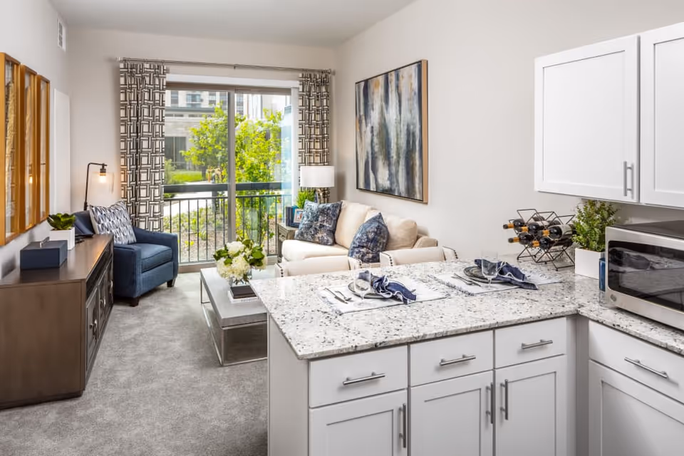 A bright and modern living area in a senior living facility featuring a small kitchen with white cabinets and granite countertops in the foreground. The kitchen counter is set with two place settings. Beyond the kitchen is a cozy living room with a beige sofa adorned with blue patterned pillows, a blue armchair, a coffee table with a flower arrangement, and a large abstract painting on the wall. Large sliding glass doors with patterned curtains open to a view of greenery outside.