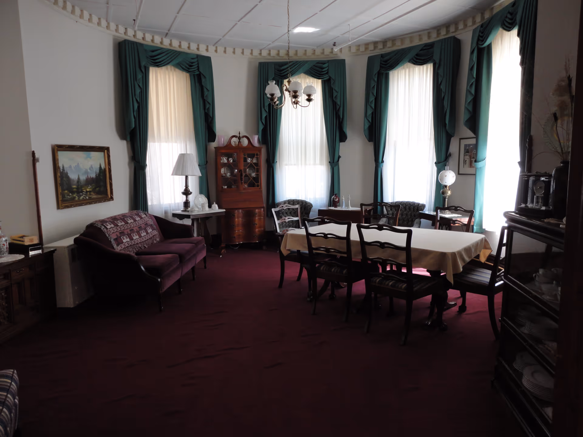 Formal dining room with a long table and chairs, a purple sofa, tall windows with green drapes, and antique furnishings.