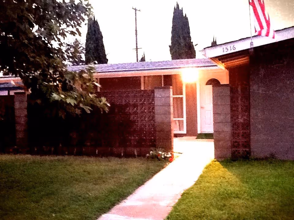 Exterior view of a single-story building with a concrete pathway leading to a white door. The building has a brick privacy wall and an American flag mounted near the roof. There are tall trees in the background and a well-maintained lawn in the foreground.