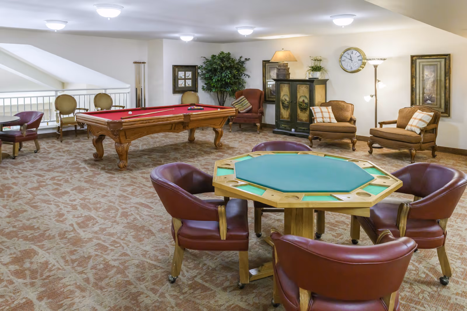 A spacious game room with a red felt pool table, an octagonal card table with four leather chairs, two armchairs with plaid pillows, a floor lamp, a cabinet, framed artwork, and a large potted plant. The room has carpeted flooring and ceiling lights.