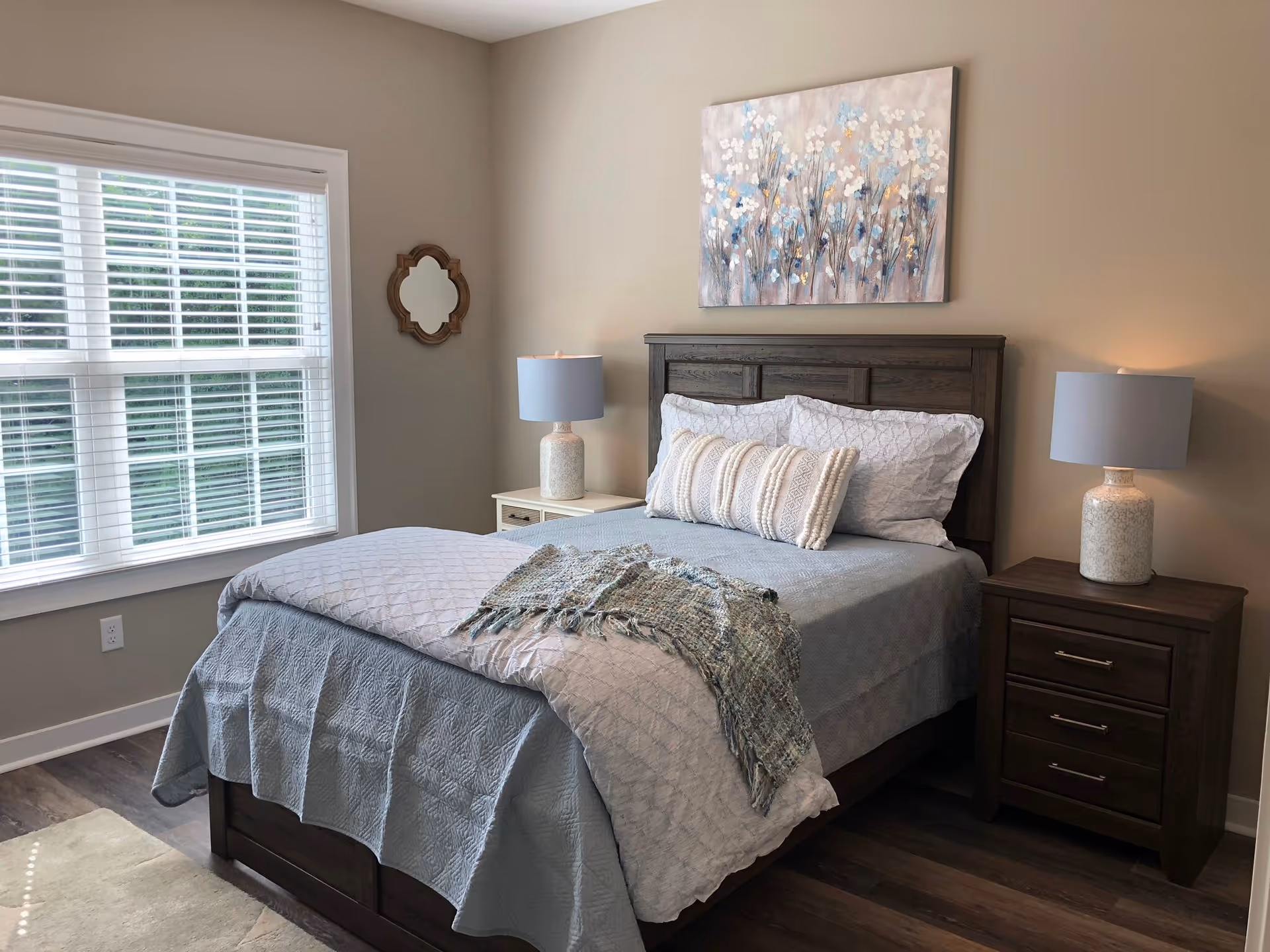 A neatly made bed with a wooden headboard in a bedroom. The bed has light blue and white bedding with a textured throw blanket and decorative pillows. On either side of the bed are matching nightstands with ceramic lamps. A window with white blinds is on the left wall, and a floral painting hangs above the bed. The room has beige walls and wood flooring.