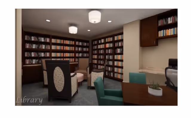 Interior view of a library room with bookshelves filled with books along the walls, several upholstered chairs arranged around a small round table, a desk with a green chair and a small plant, and ceiling lights providing illumination.