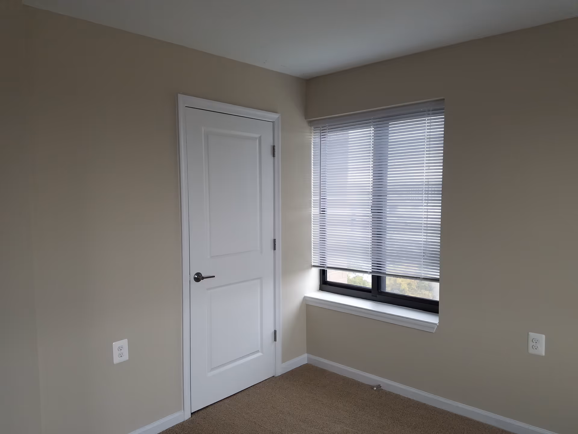 A small empty room with beige walls and carpeted floor, featuring a closed white door on the left and a window with closed blinds on the right.