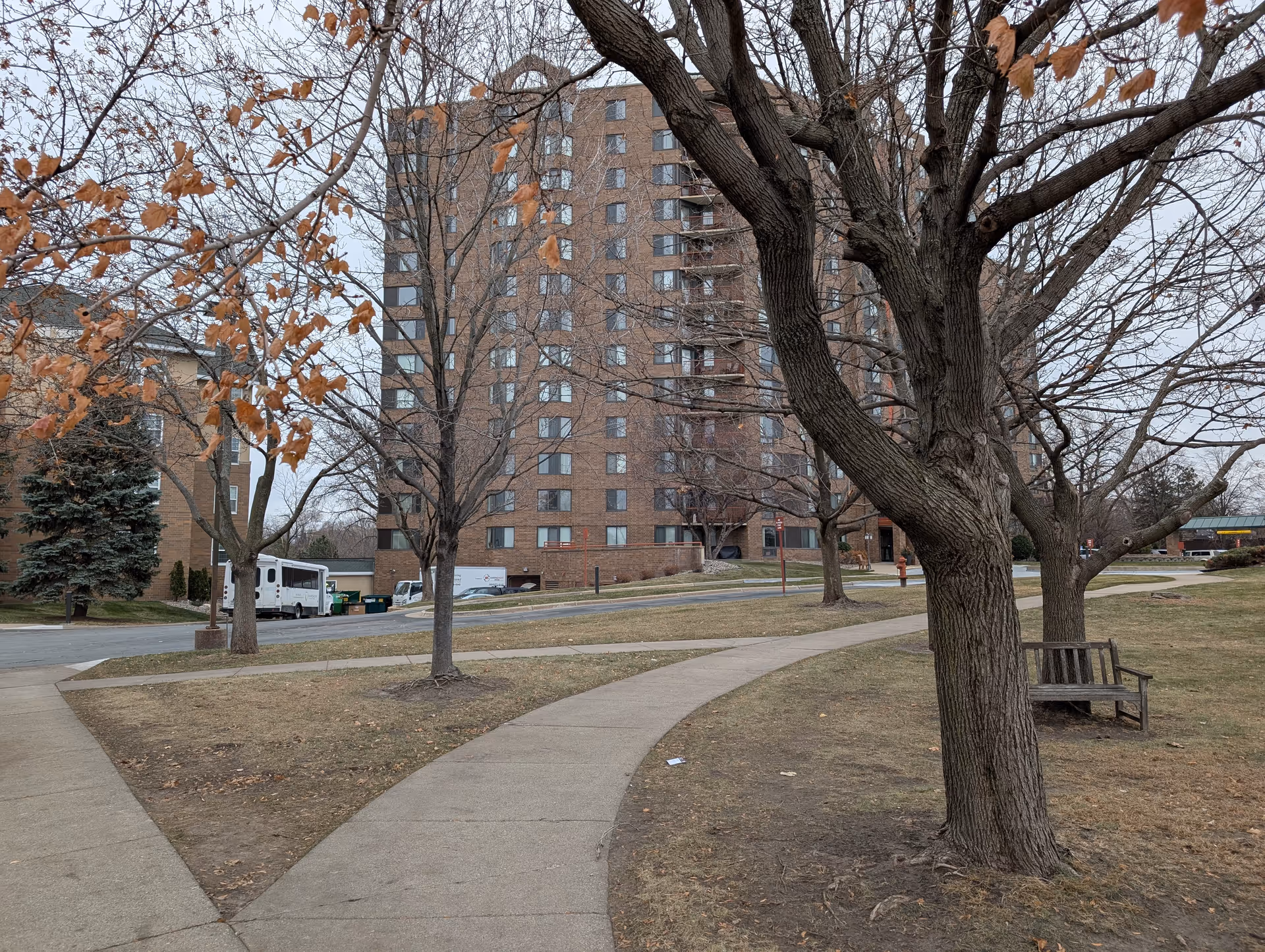Outdoor view of Parkshore Senior Campus showing a tall brick building in the background with multiple windows and balconies. In the foreground, there is a curved concrete walkway surrounded by grass and several leafless trees, one of which has a wooden bench around its base. The sky is overcast.