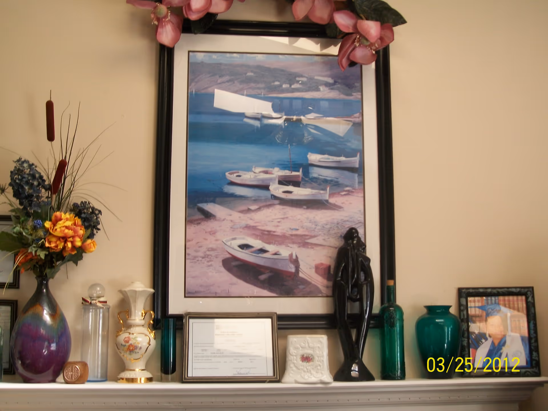A decorative shelf with various items including a large framed painting of boats on water, a black abstract sculpture, a green vase, a framed photo of a person in a graduation cap and gown, a floral arrangement in a purple vase, and several other decorative objects and framed certificates.