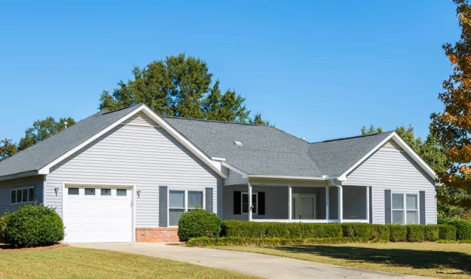 Single-story residential building with light gray siding, a dark gray shingled roof, and a white garage door. The house has a covered front porch with white columns and is surrounded by green bushes and trees under a clear blue sky.