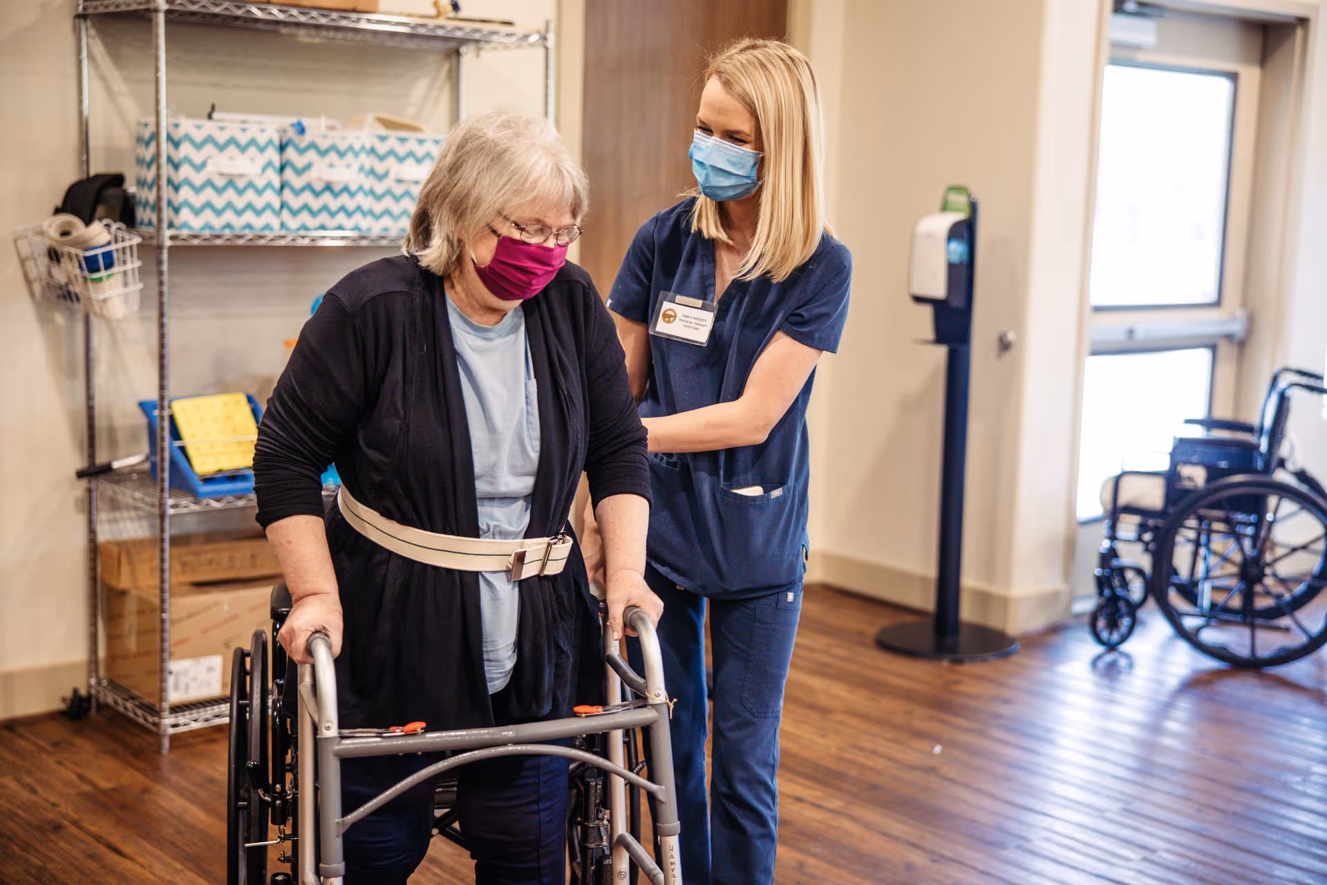 A healthcare worker wearing blue scrubs and a face mask assists an elderly woman with gray hair and a purple face mask who is using a walker in a room with wooden floors. There is a wheelchair and a hand sanitizer dispenser visible in the background.