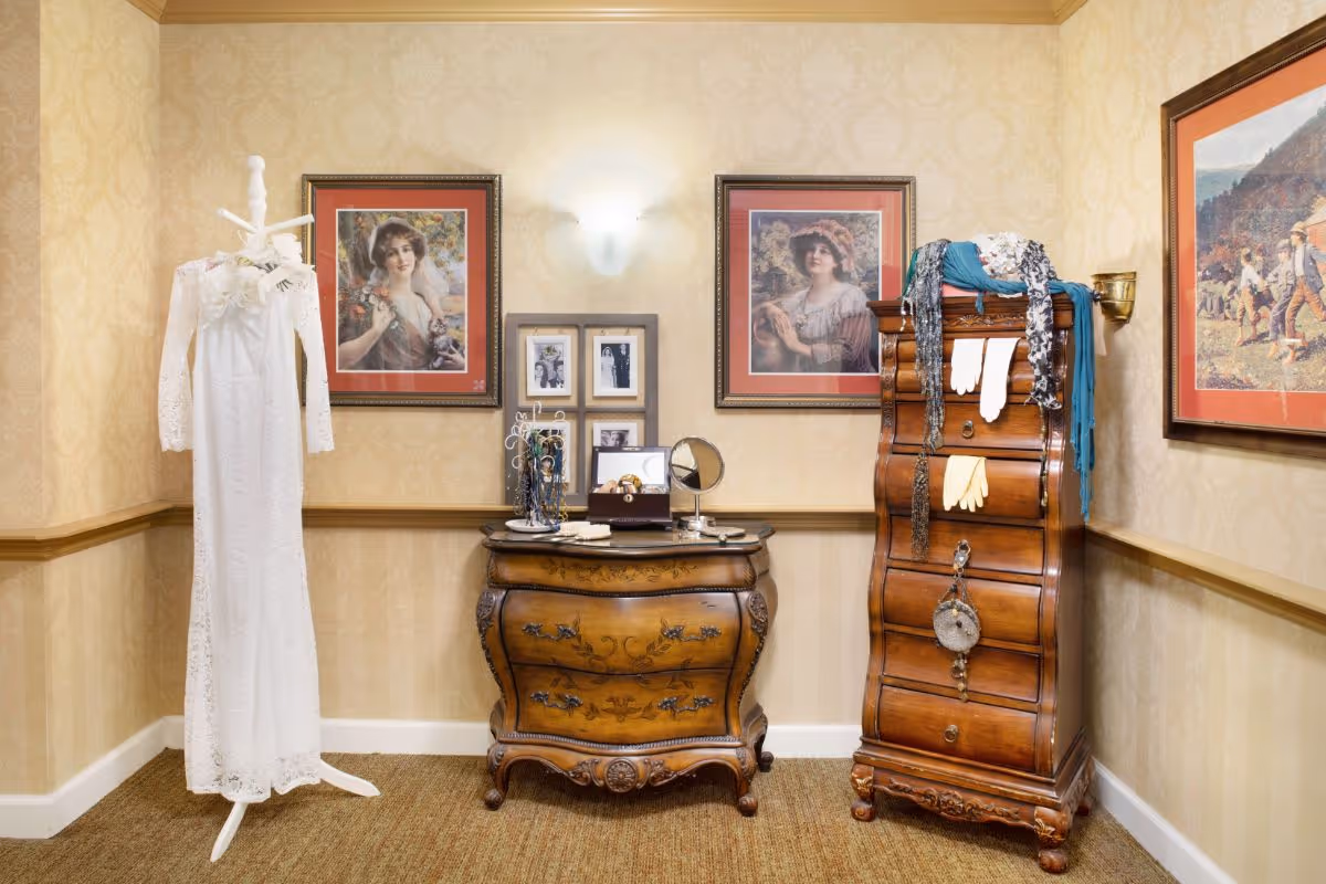 A vintage-style room with beige patterned wallpaper and carpeted floor featuring an antique wooden dresser and a tall wooden chest of drawers. A white lace dress is displayed on a white coat rack to the left. Various scarves, gloves, and jewelry are draped over the furniture. The walls are decorated with framed portraits and pictures.