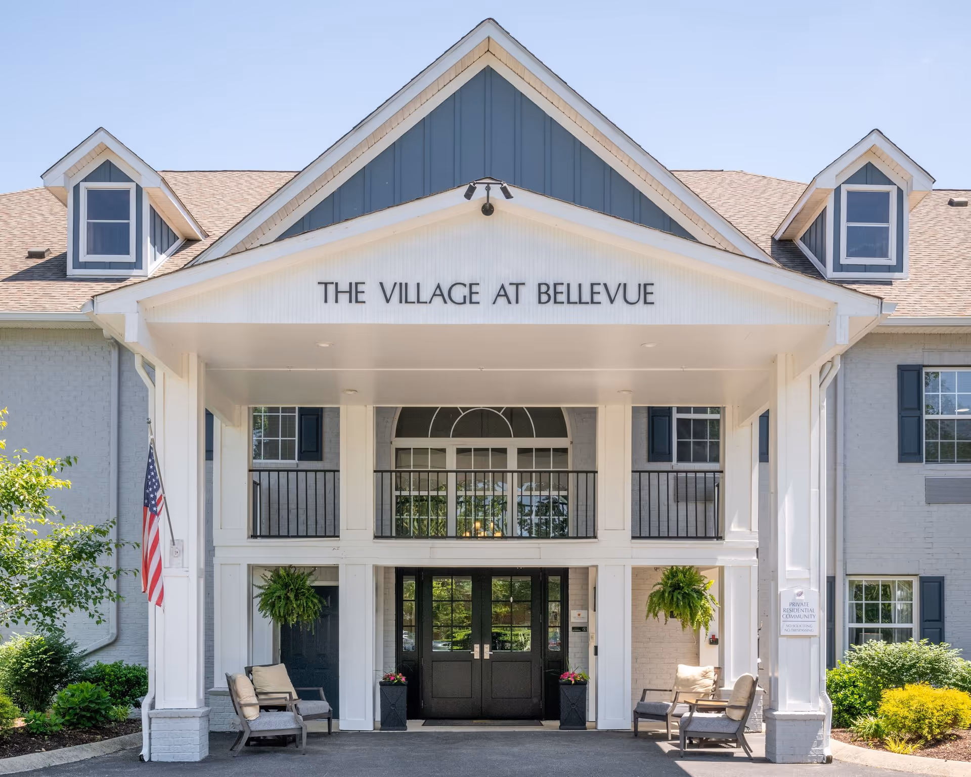 Front entrance of The Village at Bellevue assisted living facility with a covered porch, seating area with chairs, hanging plants, and an American flag on the left side.