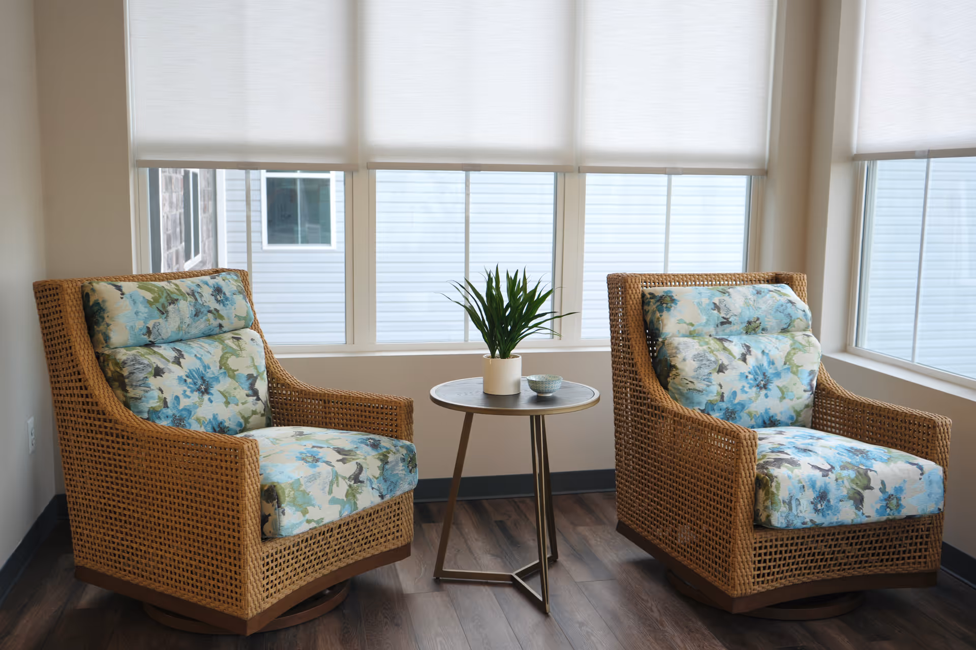 Two wicker armchairs with blue floral cushions flank a small round side table with a potted plant in a bright seating area by windows.