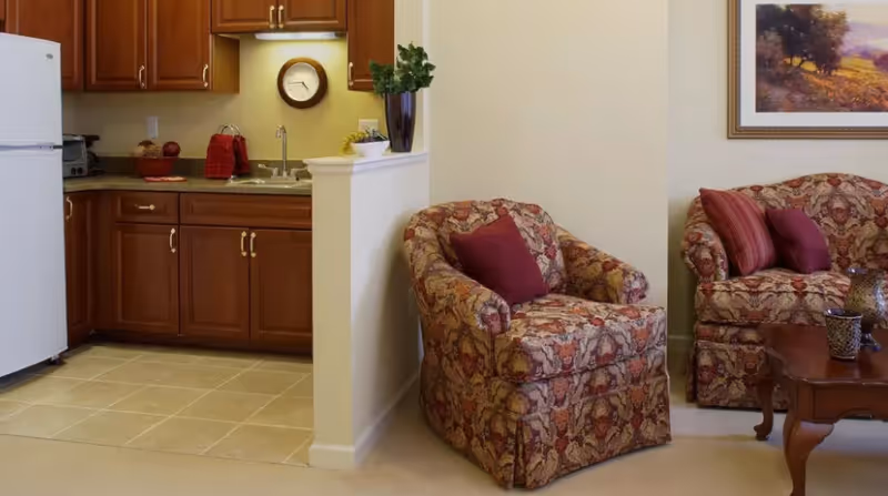 Open living area with patterned armchairs and a matching sofa beside a small kitchen with wooden cabinets and a refrigerator.