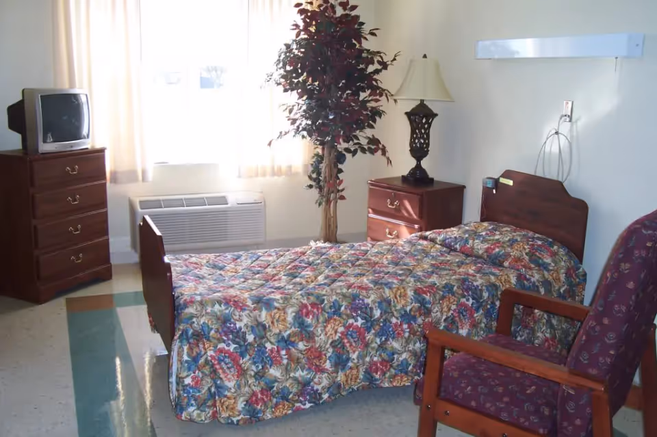 A small bedroom in a senior living facility with a single bed covered in a floral bedspread, a wooden nightstand with a decorative lamp, a wooden dresser with an old-style TV on top, a potted artificial tree, and a cushioned armchair with wooden armrests. The room has a window with light curtains and a wall-mounted air conditioning unit beneath it.