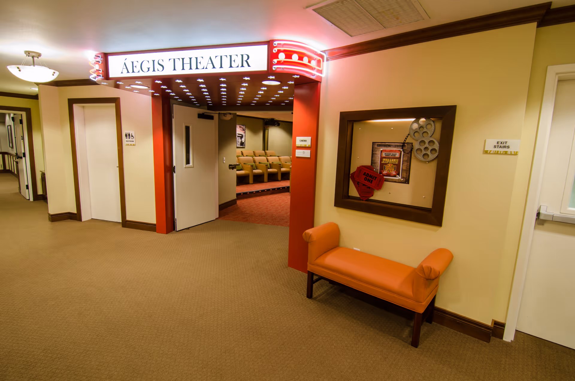 Interior hallway of a senior living facility leading to the Aegis Theater. The theater entrance has a lit marquee sign with the name 'Aegis Theater' and red decorative lights. Inside, rows of beige theater seats are visible. To the right of the entrance is an orange bench and a framed display with movie-related items, including a film reel and 'Admit One' tickets. There are doors labeled for exit stairs and restrooms along the hallway.