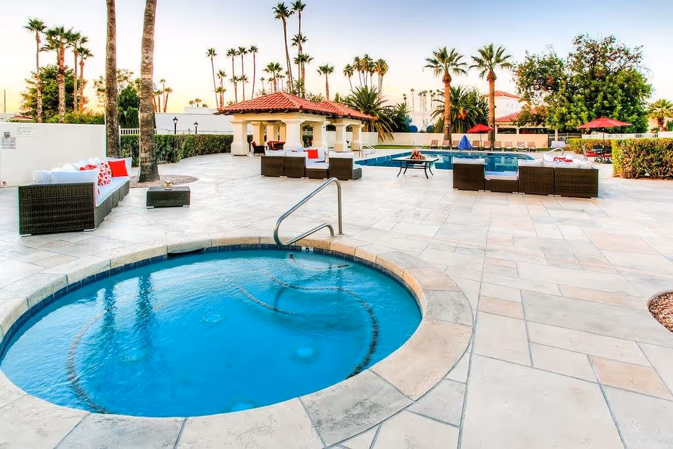 Outdoor area at Independent Living at Arizona Golf Resort featuring a circular hot tub in the foreground, a swimming pool with lounge chairs and umbrellas, palm trees, and a covered seating area with a red-tiled roof.
