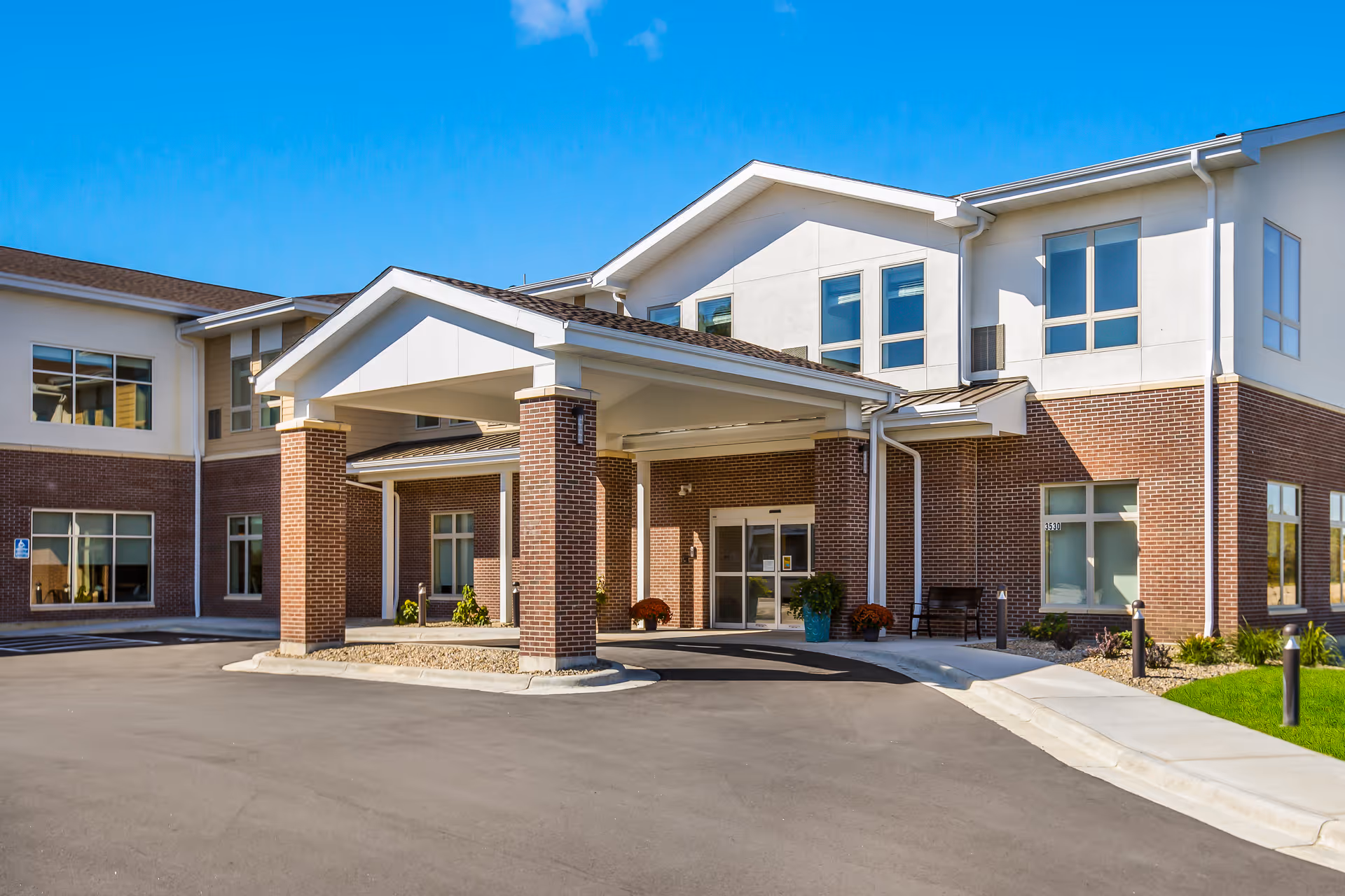 Exterior view of a modern senior living facility building with a covered entrance supported by brick columns, large windows, and a clear blue sky.