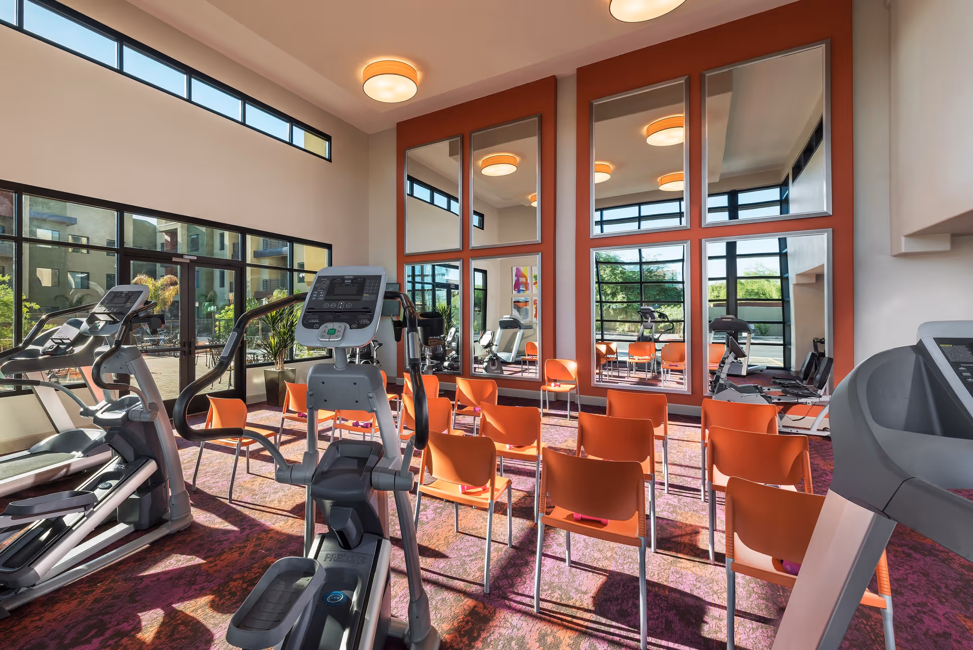 A bright fitness room with large windows and mirrors on the wall. The room contains several orange chairs arranged in rows and various exercise machines including treadmills and elliptical trainers. The floor has a patterned carpet and the walls are painted in neutral tones with an accent wall in orange.