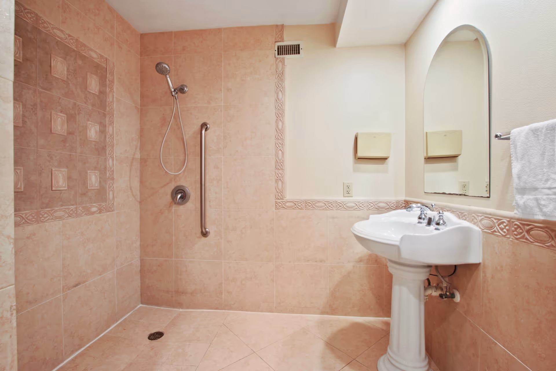 A bathroom with beige tiled walls and floor featuring a walk-in shower with a handheld showerhead and a grab bar. There is a white pedestal sink with a mirror above it and a towel rack with a white towel on the right side.