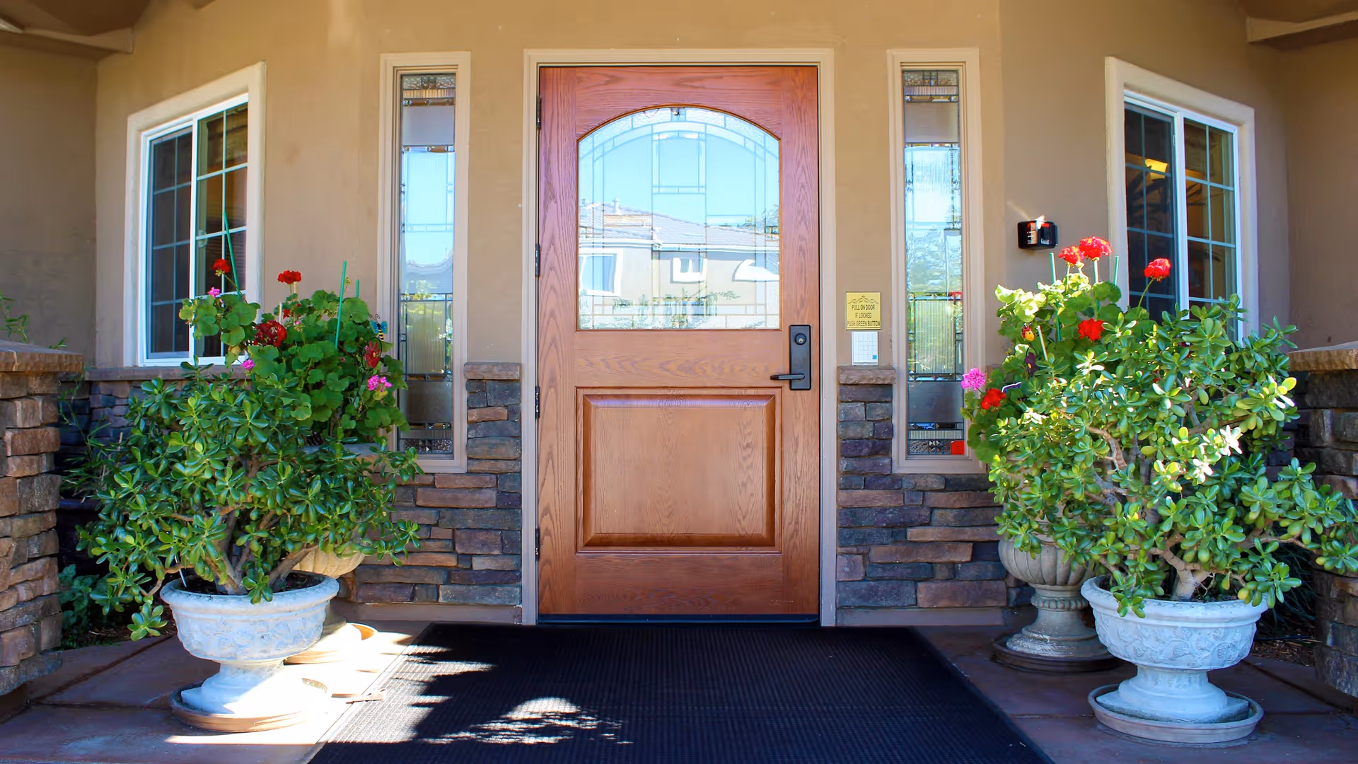 Front entrance of a building with a wooden door featuring decorative glass panels on either side and above. Two large white planters with green plants and red flowers flank the door. The exterior walls are beige with stone accents around the door and windows.