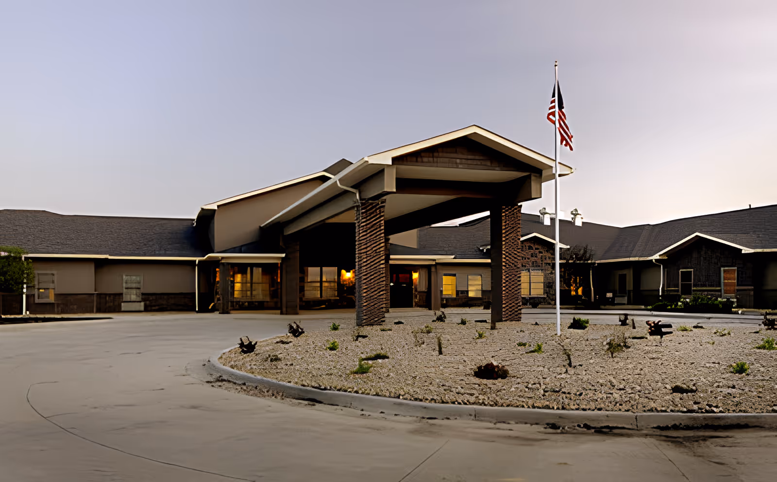 Exterior view of Carriage Crossing Senior Living of Champaign at dusk, showing the building entrance with a covered drop-off area supported by brick pillars, a circular driveway, and an American flag on a flagpole in a landscaped area with rocks and small plants.