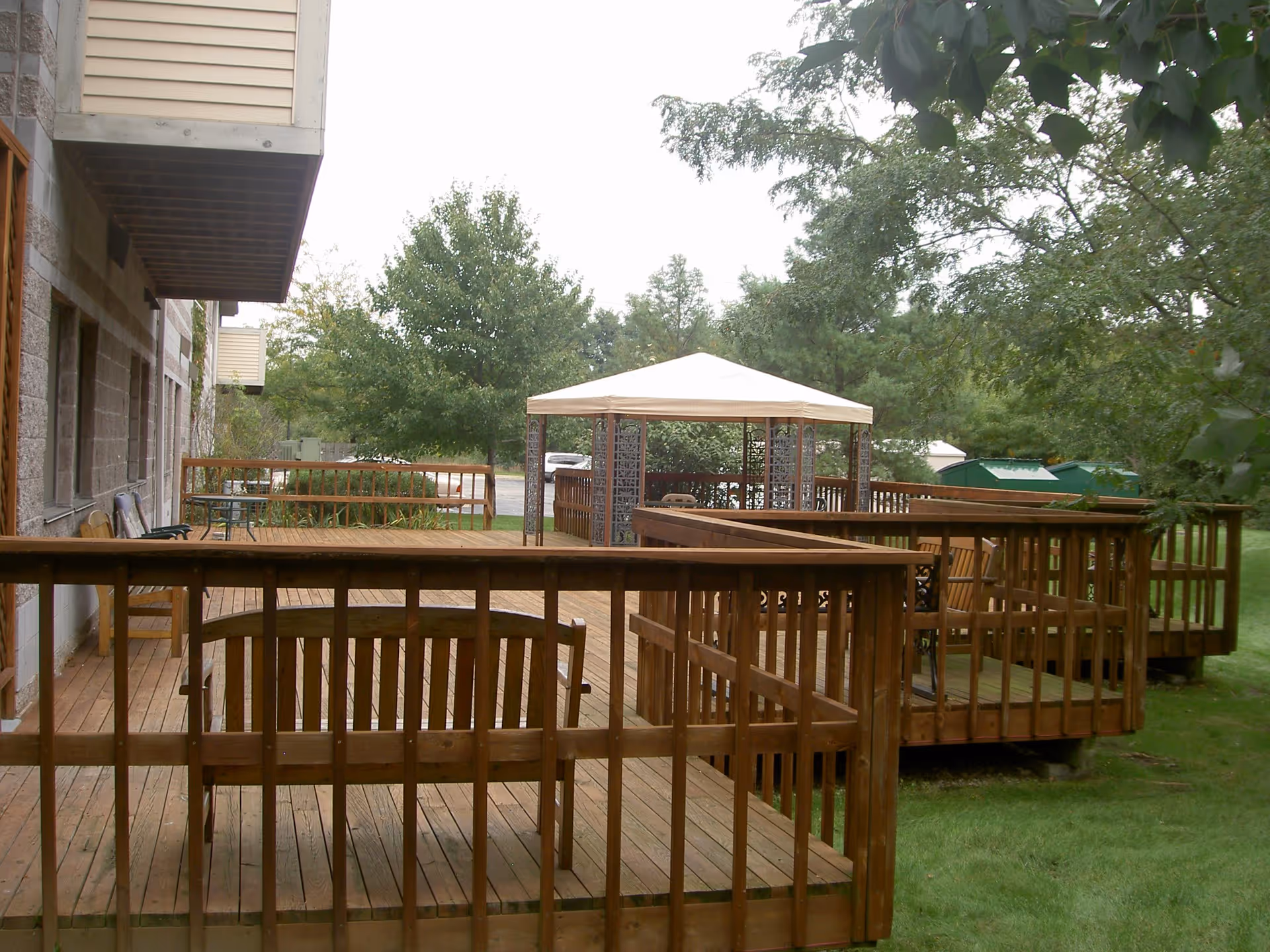 Outdoor wooden deck area attached to a building with multiple seating areas and a gazebo with a white canopy, surrounded by green grass and trees.