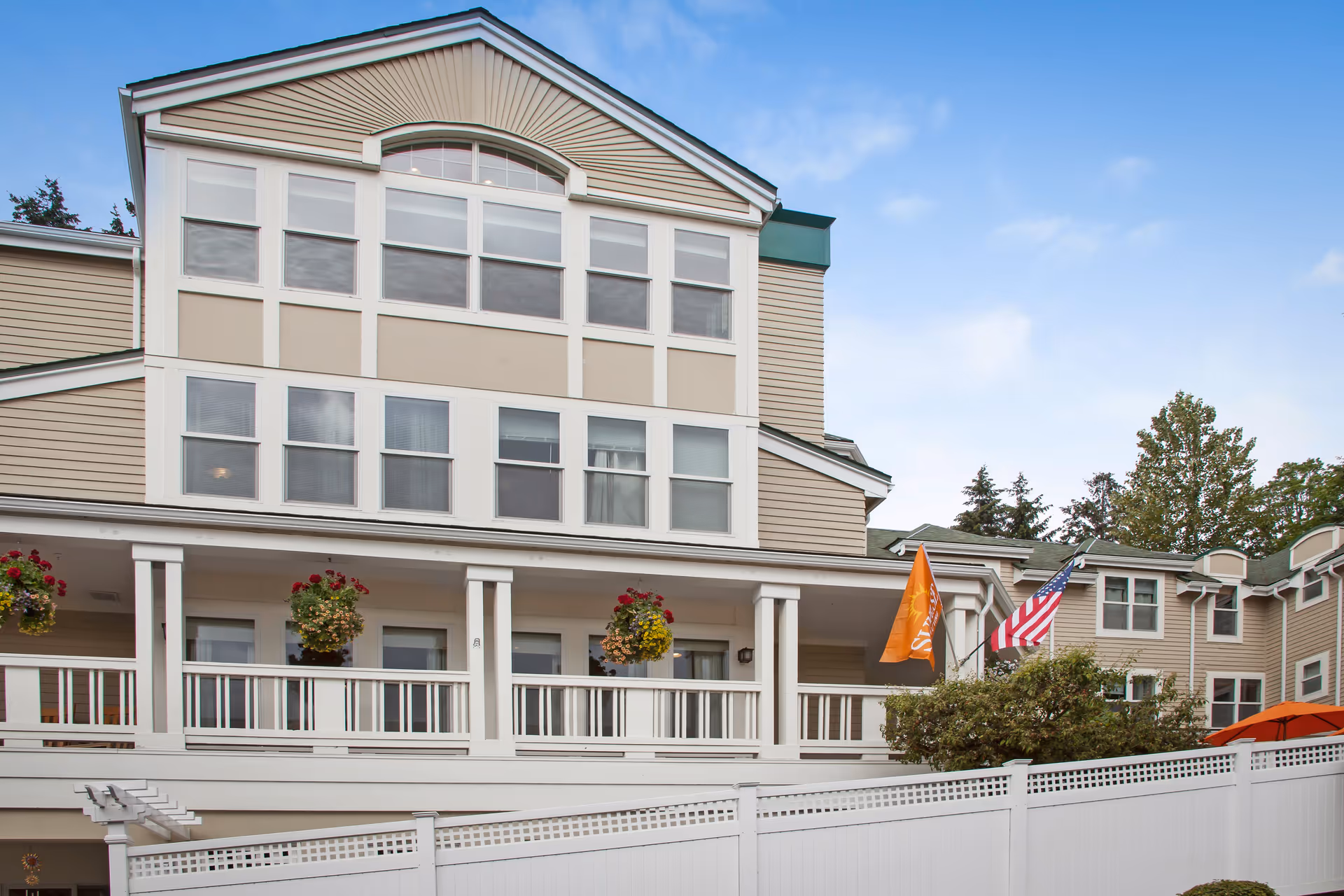 Exterior view of a multi-story senior living facility building with beige siding, multiple windows, a white balcony with hanging flower baskets, and two flags including an American flag. There is a white fence in the foreground and some trees in the background under a partly cloudy sky.